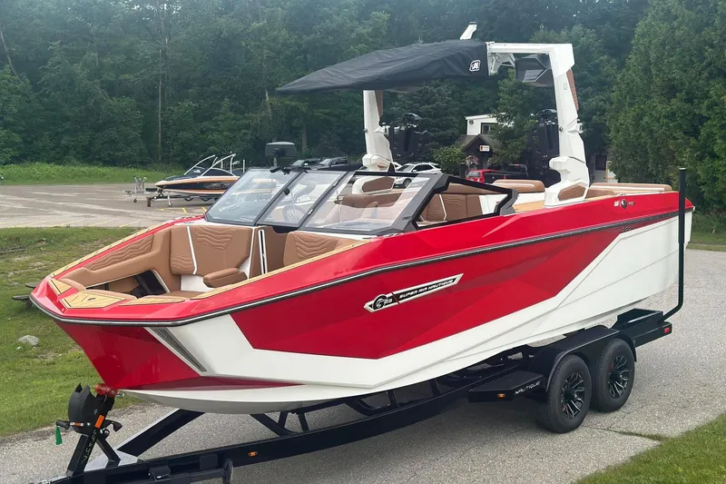 Slide: The Image of 2025 Nautique G23 boat in red and white, parked on a trailer, surrounded by greenery. - 2