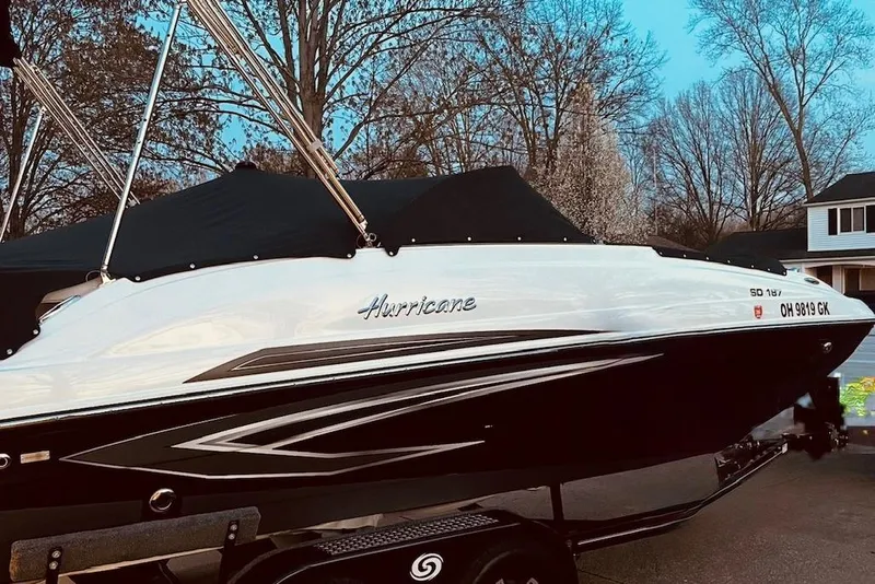 Slide: The Image of 2022 Hurricane SunDeck 187 IO boat on trailer, parked outdoors with trees in background. - 2