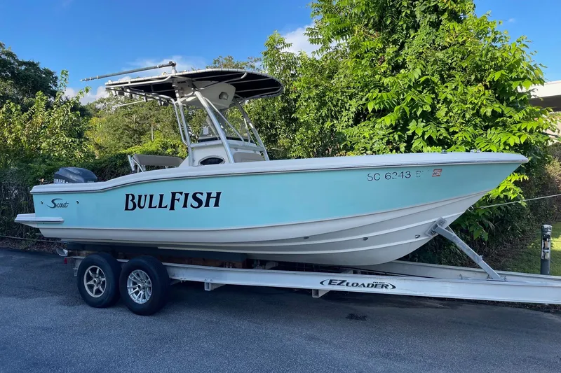 Slide: The Image of 2006 Scout 242 Sportfish boat on trailer, labeled "Bullfish," parked near lush greenery. - 5