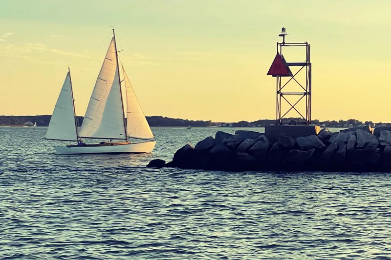 Slide: The Image of 1974 Herreshoff Rozinante sailboat near a rocky shoreline at sunset. - 4