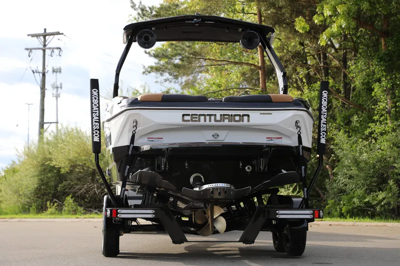 Slide: The Image of 2025 Centurion Nv213 boat rear view, parked on road with lush greenery. - 25