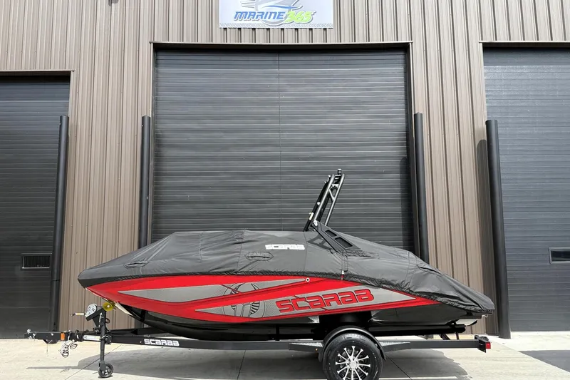 Slide: The Image of 2026 Scarab 195 LX boat covered, parked outside a garage with a black and red design. - 32