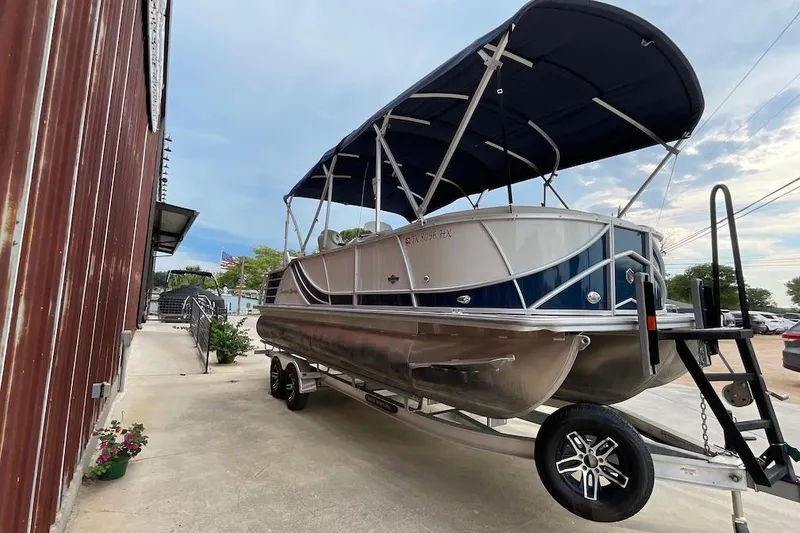 Slide: The Image of 2020 South Bay 523 pontoon boat on trailer beside a rustic building. - 2