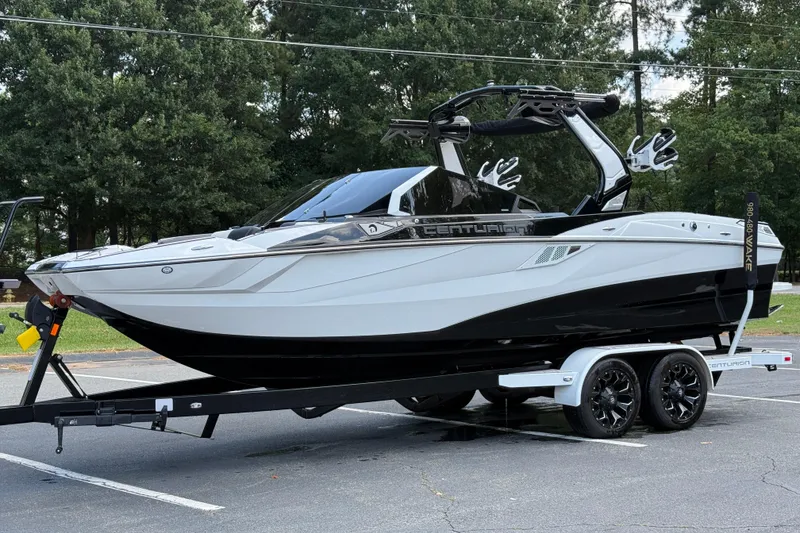 Slide: The Image of 2024 Centurion Fi23 boat on trailer, parked in a lot with trees in the background. - 15