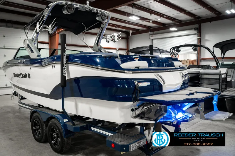 Slide: The Image of 2025 MasterCraft X24 boat in showroom, blue and white design, on trailer with illuminated platform. - 10