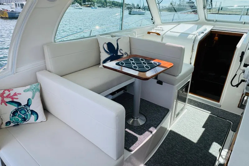 Slide: The Image of 2025 Back Cove 34O boat interior with plush seating, nautical-themed pillows, and a wooden table. - 21