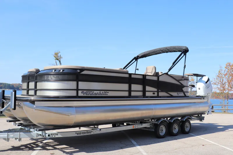 Slide: The Image of 2023 Vanderbilt 700 Twin pontoon boat on trailer, parked by a lake. - 0