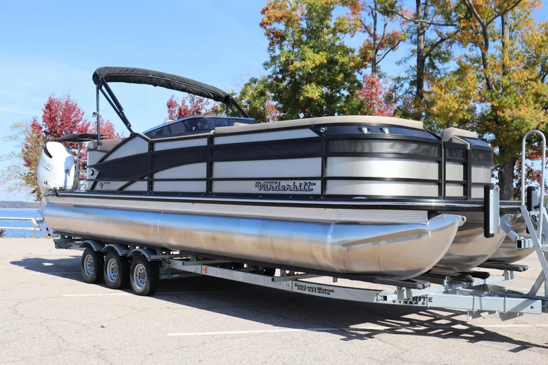 Slide: The Image of 2023 Vanderbilt 700 Twin pontoon boat on trailer, parked near autumn trees. - 14