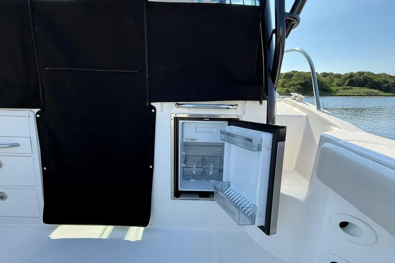 Slide: The Image of 2026 Boston Whaler 285 Conquest boat interior with open refrigerator, docked by a scenic lake. - 19