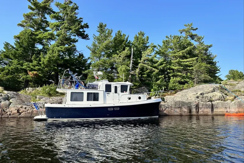 Slide: The Image of 2005 American Tug 34 Pilothouse boat anchored near rocky shoreline with trees. - 126