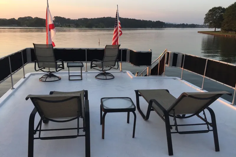 Slide: The Image of 1998 Sumerset 16x80 houseboat deck with chairs, tables, and flags overlooking serene lake at sunset. - 12
