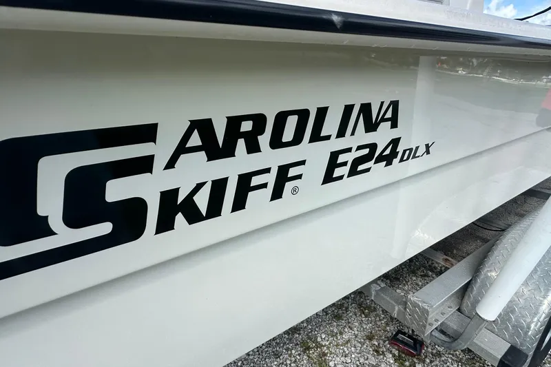 Slide: The Image of 2024 Carolina Skiff E24 DLX boat logo on white hull, parked on trailer. - 13