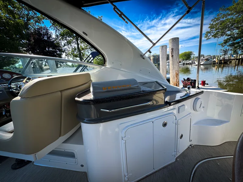Slide: The Image of 2016 Rinker 290 Express boat interior with seating and outdoor kitchen, docked by a scenic waterfront. - 26