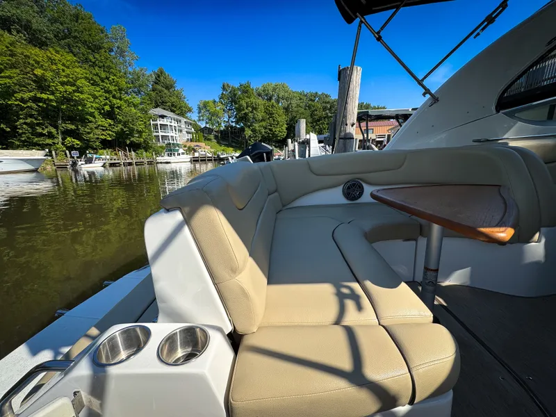 Slide: The Image of 2016 Rinker 290 Express boat interior with beige seating and wooden table, docked by a scenic shoreline. - 35