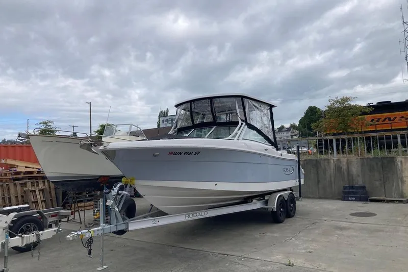 Slide: The Image of 2021 Robalo R247 Dual Console boat on trailer, parked outdoors under cloudy sky. - 2