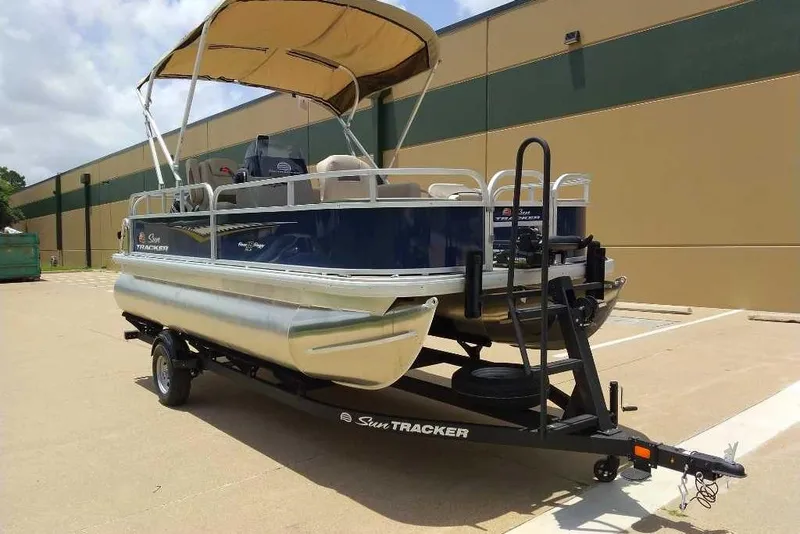 Slide: The Image of 2025 Sun Tracker Bass Buggy 18 DLX pontoon boat on trailer, parked outdoors. - 2