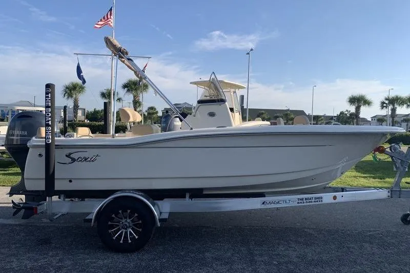 Slide: The Image of 2026 Scout 195 Sportfish boat on trailer, parked outdoors with flags and palm trees. - 4