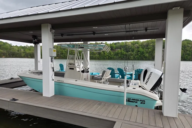 Slide: The Image of 2025 Carolina Skiff 23 Ultra Elite boat docked on a serene lake under a covered pier. - 2