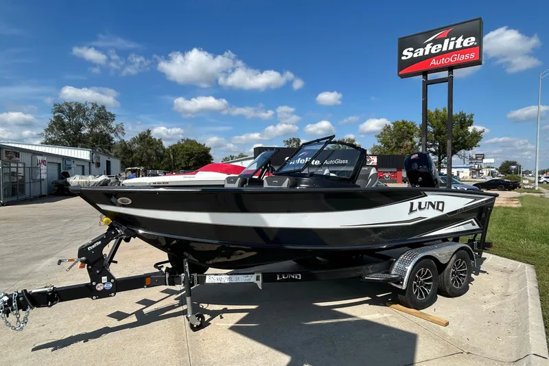 Slide: The Image of 2026 Lund 1875 Crossover XS boat on trailer, parked near Safelite AutoGlass sign. - 2
