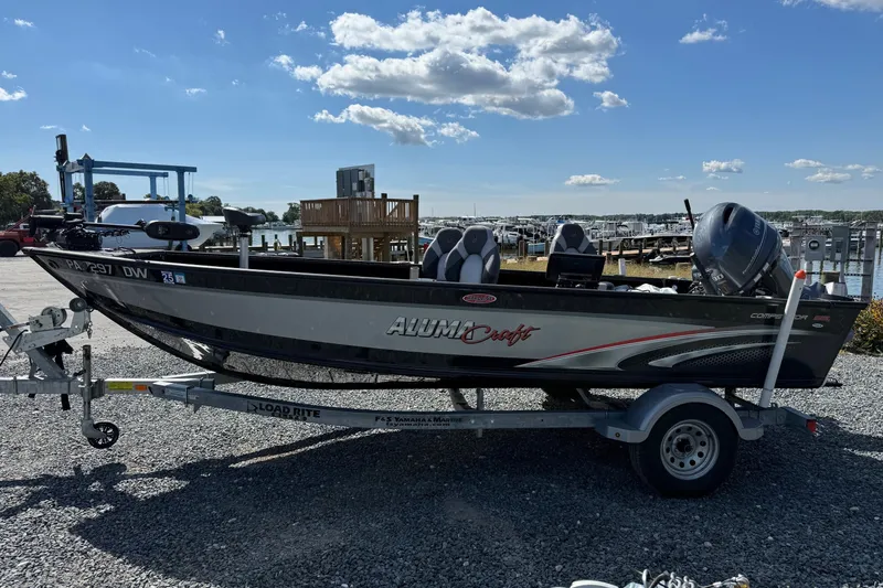 Slide: The Image of 2019 Alumacraft Competitor 185 CS boat on trailer, parked near marina under blue sky. - 6