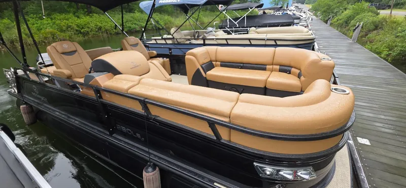 Slide: The Image of 2024 Regency 230 LE3 Sport pontoon boat with tan seating, docked by a lush green shoreline. - 15