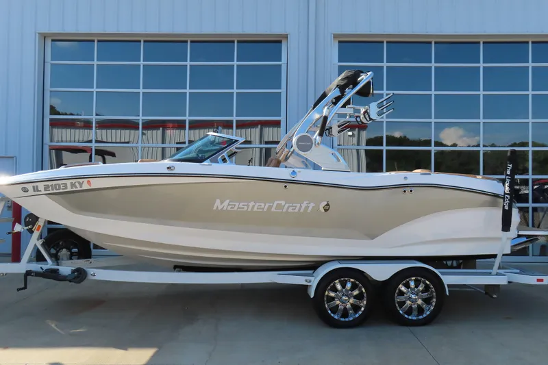 Slide: The Image of 2019 MasterCraft X22 boat on trailer, parked outside a building with large windows. - 0