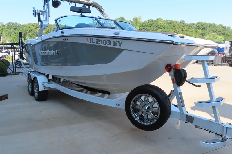 Slide: The Image of 2019 MasterCraft X22 boat on trailer, parked outdoors, with lush green trees in background. - 2
