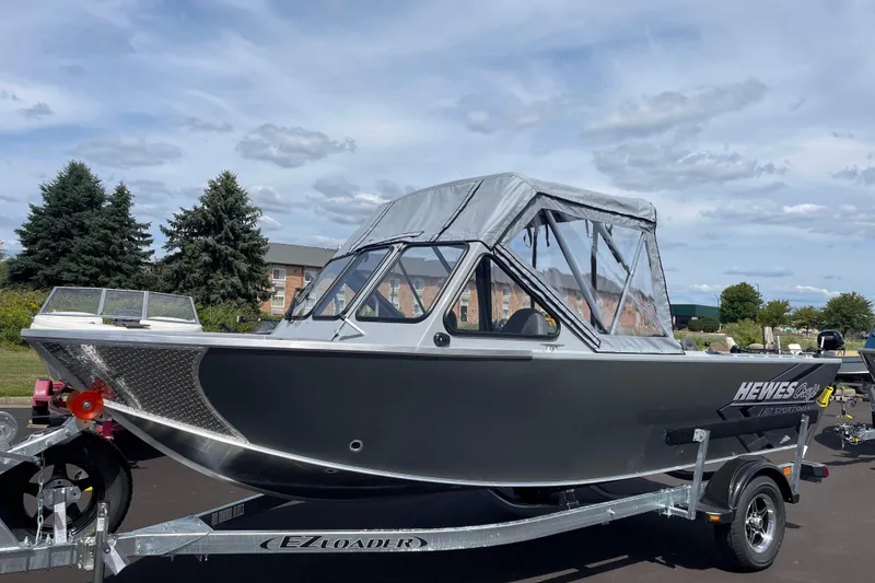The Image of 2026 Hewescraft 180 Sportsman boat on trailer, parked outdoors under cloudy sky. - 0