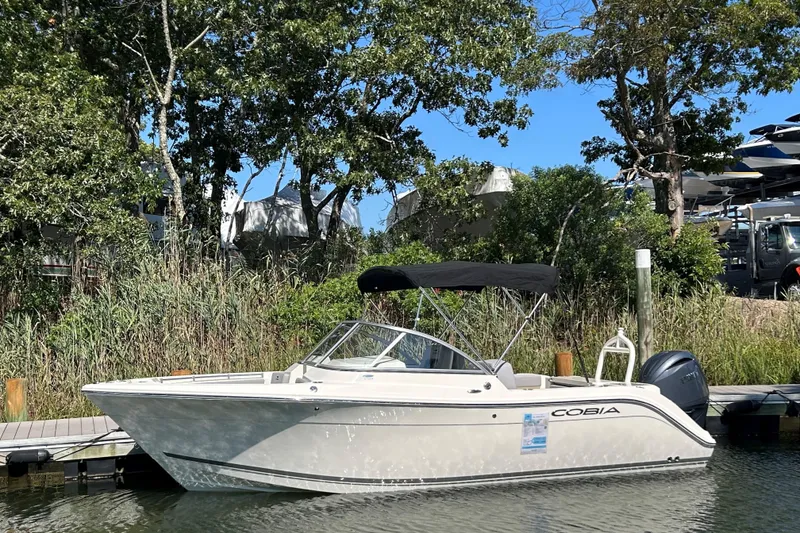Slide: The Image of 2026 Cobia 220 Dual Console boat docked by lush greenery. - 1