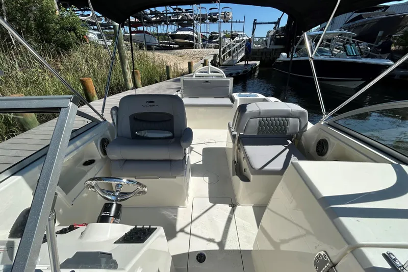 Slide: The Image of 2026 Cobia 220 Dual Console boat interior with seating and steering wheel, docked by the water. - 14