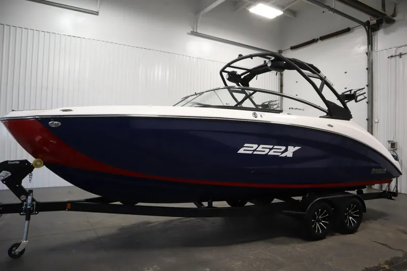 Slide: The Image of 2025 Yamaha Boats 252XE in showroom, sleek design, blue and red hull, on trailer. - 7