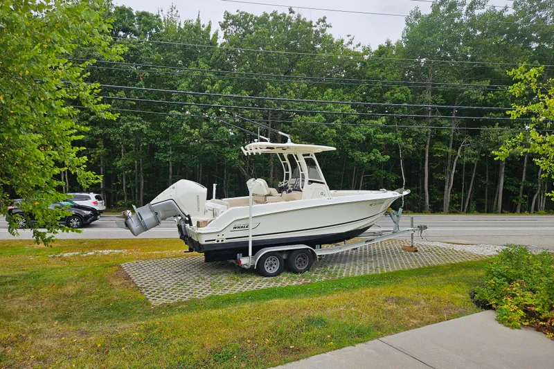 Slide: The Image of 2019 Boston Whaler 230 Outrage boat on trailer, parked near a road with trees in the background. - 20