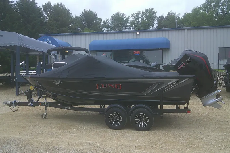 Slide: The Image of 2025 Lund 1875 Pro-V boat with cover, parked on a trailer outside a building. - 21