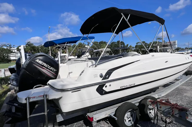 The Image of 2018 Bayliner 210 Deck Boat on trailer with Mercury outboard motor, under blue sky. - 1