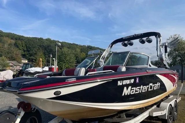 The Image of 2013 MasterCraft X-25 boat on trailer by a lake under a clear sky. - 0