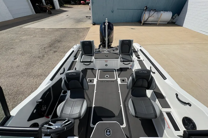 Slide: The Image of 2026 Lund 185 Impact GL boat interior with seating and steering wheel, parked outdoors. - 16