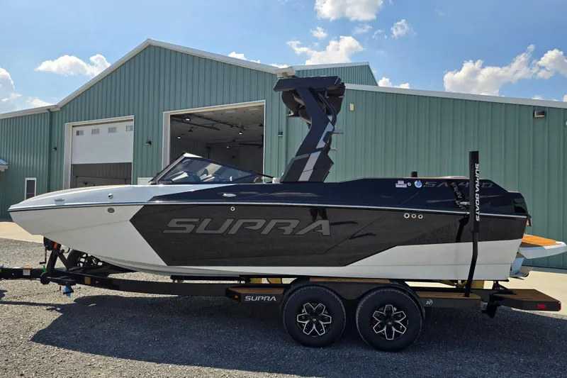 The Image of 2026 Supra SA boat on trailer, parked outside a large green building under a blue sky. - 1