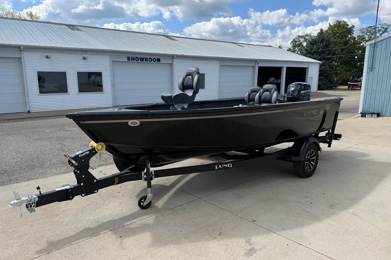 Slide: The Image of 2026 Lund 1800 Explorer Tiller boat on trailer outside showroom, under cloudy sky. - 3