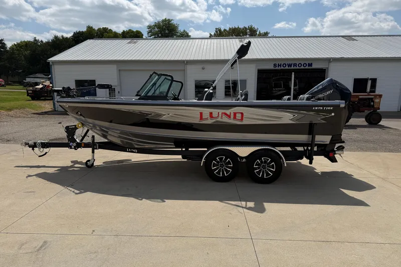 Slide: The Image of 2026 Lund 1975 Tyee boat on trailer outside showroom under cloudy sky. - 4