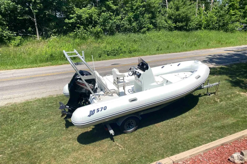 Slide: The Image of 2013 Mercury Inflatables M570 boat on trailer, parked on grass by a road. - 2