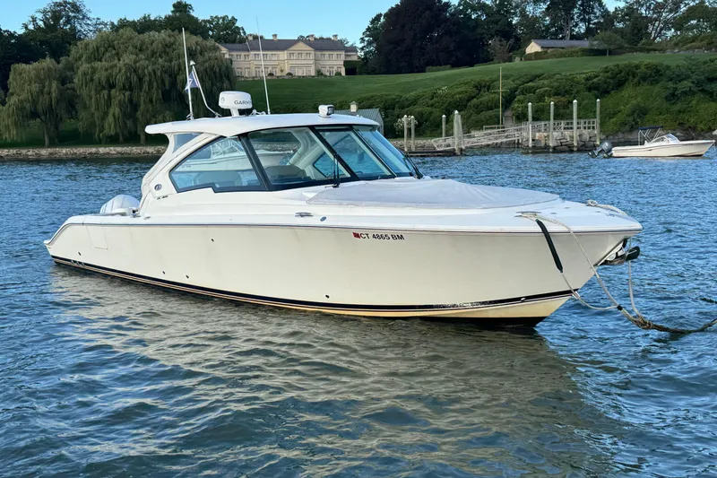 Slide: The Image of 2021 Pursuit DC 326 Dual Console boat on calm water near a lush shoreline. - 2