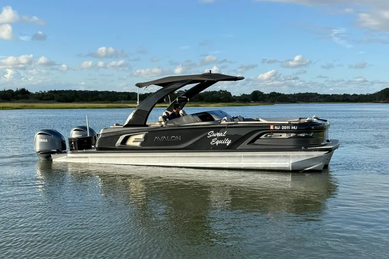 Slide: The Image of 2025 Avalon Excalibur Quad Lounge Windshield boat on calm water under blue sky. - 8
