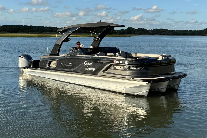 Slide: The Image of 2025 Avalon Excalibur Quad Lounge Windshield boat on calm water under blue sky. - 9