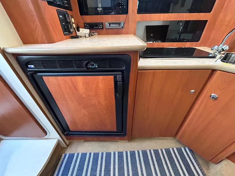 Slide: The Image of 2007 Bayliner 305 boat kitchen with wood cabinetry, fridge, sink, and striped rug. - 24