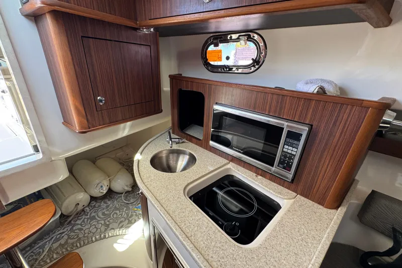 Slide: The Image of 2015 Pursuit OS 285 Offshore boat interior with kitchenette, microwave, sink, and wooden cabinetry. - 28
