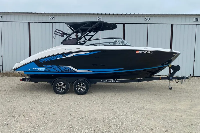 The Image of 2021 Yamaha Boats 252XE on trailer, black and blue design, parked outside storage units. - 0