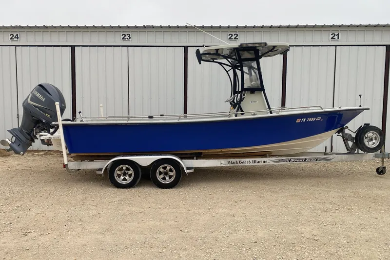 The Image of 2015 Blazer Bay 2400 boat on trailer, blue hull, Yamaha outboard motor, parked by storage units. - 0
