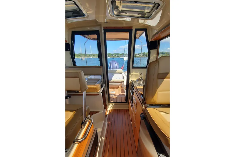 Slide: The Image of 2024 Ranger Tugs R-27 interior with tan seating, wood flooring, and scenic water view. - 21