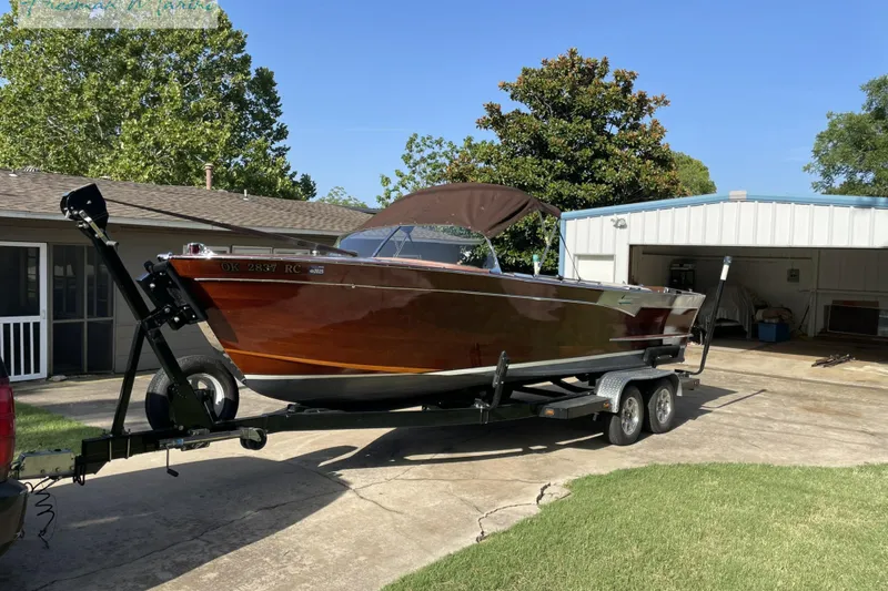 The Image of 1964 Shepherd 22 boat on trailer, parked in driveway, surrounded by trees and buildings. - 0