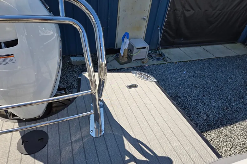 Slide: The Image of 2025 Godfrey Monaco 255 SB boat deck with railing and engine, parked on gravel. - 28